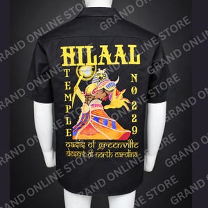 May include: A black short-sleeved button-down shirt featuring a large yellow graphic on the back. The design includes the text HILAAL, TEMPLE, NO 229, and OASIS OF GREENVILLE DESERT OF NORTH CAROLINA, alongside an illustration of an Egyptian deity.