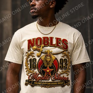 May include: A cream-coloured t-shirt featuring a graphic print with the text NOBLES, 1893, and Mystic Shrine. The design includes a gold pharaoh mask, a red star, a curved sword, and the text ANCIENT EGYPTIAN ARABIC ORDER in a decorative gold frame.