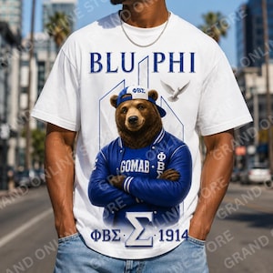 May include: A white short-sleeve t-shirt featuring a graphic of a brown bear wearing a blue baseball cap and a blue varsity jacket. The text BLU PHI, GOMAB, and the Greek letters ΦΒΣ and Σ appear alongside the year 1914 and a flying white dove.