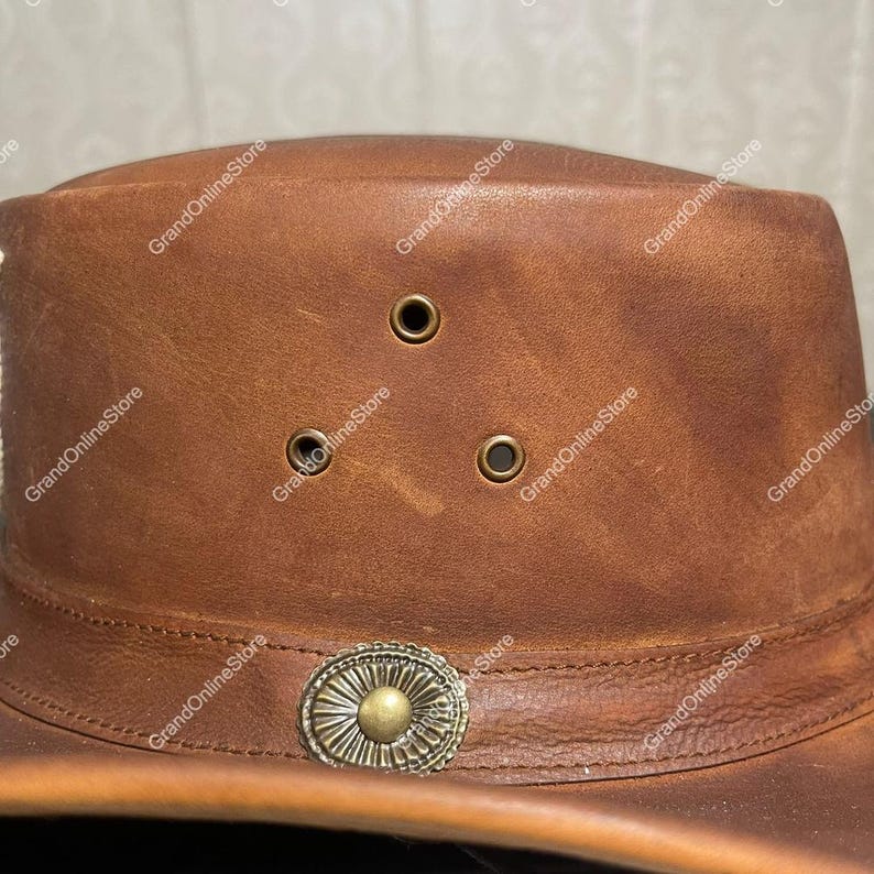 May include: A brown leather hat with a wide brim. The hat has three ventilation eyelets and a decorative metal band around the crown. The hat has a distressed finish, giving it a vintage appearance.