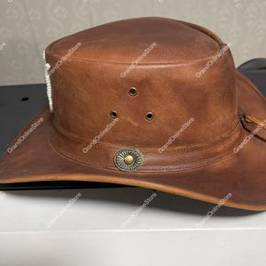 May include: A brown leather cowboy hat with a wide brim. The hat has three small air vents, a decorative metal emblem, and a white beaded accent. The hat is designed with a classic western style.
