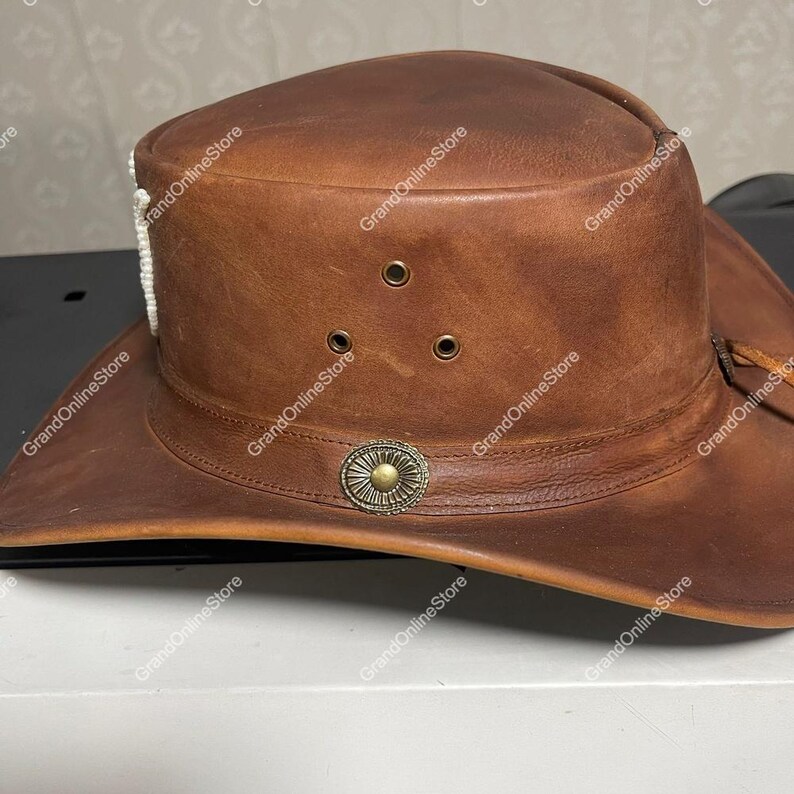May include: A brown leather cowboy hat with a wide brim. The hat has three small air vents, a decorative metal emblem, and a white beaded accent. The hat is designed with a classic western style.
