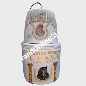 May include: White ceremonial hat and matching cylindrical case. The hat features the word "CHAM" in jeweled letters and a jeweled emblem. The case is embroidered with text and an emblem. A black handle is attached to the top of the hat.