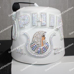 May include: White fez hat with a black tassel and a rhinestone design. The fez has the word "PHILA" spelled out in rhinestones and a small image of an Egyptian pharaoh.
