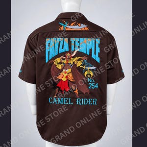 May include: A brown short-sleeve button-down shirt featuring a large back graphic of a warrior figure, a sword, and the text FAYZA TEMPLE CAMEL RIDER No. 254 in light blue. The design includes a smaller sword graphic above the collar area.