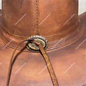 May include: Close-up of a brown leather cowboy hat. The hat has a decorative metal band around the crown and a leather chin strap. The leather has a distressed appearance, with visible stitching and a worn texture. A classic Western style hat.