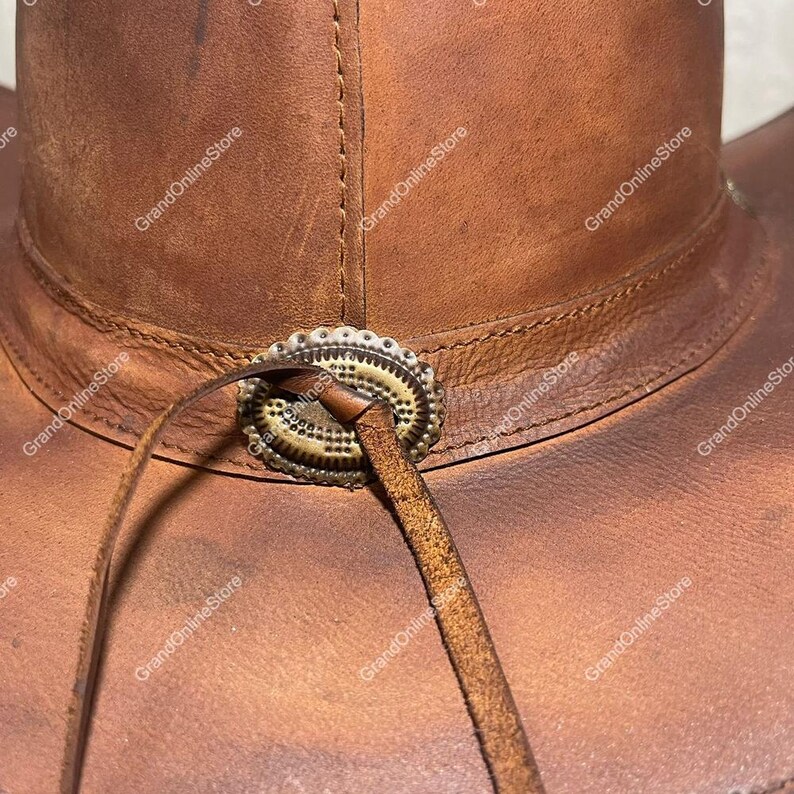 May include: Close-up of a brown leather cowboy hat. The hat has a decorative metal band around the crown and a leather chin strap. The leather has a distressed appearance, with visible stitching and a worn texture. A classic Western style hat.