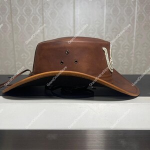 May include: A brown leather hat with a wide brim and a decorative white beaded accent. The hat has a structured crown with three eyelets and a chin strap. The hat's brim is approximately 7.6 cm wide.