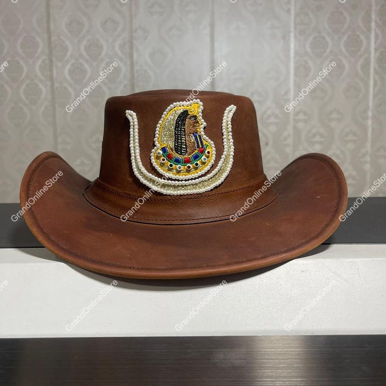May include: A brown leather cowboy hat with an Egyptian-themed emblem. The emblem is decorated with gold, pearls, and colourful details. The hat has a wide brim and a traditional cowboy hat silhouette. The hat's measurements are in centimeters.