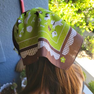 May include: A square headscarf featuring a lime green center with a repeating pattern of white dogs, bones, and yarn balls. The border is dark brown with a white scalloped fence design and small lime green flower accents at the corners.
