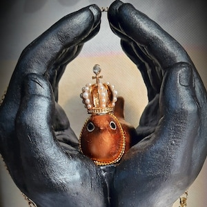 May include: A small, brown bird figurine wearing a gold crown with white pearls. The bird is held in two black hands, creating a protective gesture.