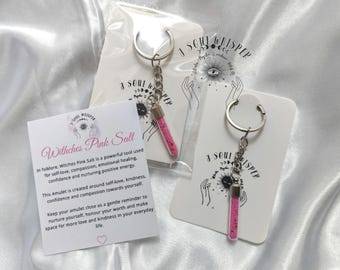 May include: Two keychains featuring small, clear glass vials filled with pink salt, attached to silver-toned metal rings. Each is mounted on a white card labeled A Soul Whisper. A square card titled Witches Pink Salt provides descriptive text.