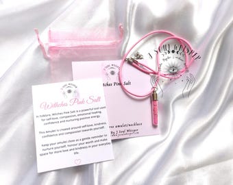 May include: A pink cord necklace featuring a small glass vial pendant filled with pink salt. The set includes a pink organza drawstring pouch and a white informational card with the text Witches Pink Salt and J Soul Whisper branding.