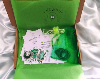 May include: A cardboard box lined with green tissue paper containing white informational cards, a green organza drawstring bag, a coil of green string, and two stickers featuring a green crescent moon, an eye, and a crystal illustration.