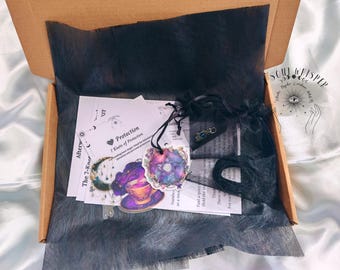 May include: A cardboard box containing black tissue paper, white informational cards with text including Protection and 7 Knots of Protection, a small black drawstring bag, a black cord, and colorful stickers featuring a pentagram and a teacup.