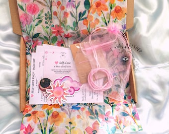 May include: A cardboard box lined with floral tissue paper containing a Self-Love 6 Knots of Self-Love magic kit. The kit includes a pink string in a sheer organza bag, a sun-shaped sticker, a cloud-shaped sticker, and an instructional card.