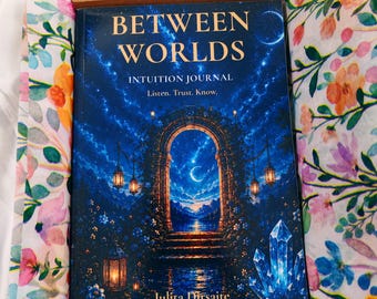 May include: A journal titled Between Worlds Intuition Journal by Julita Dirsaite, featuring a dark blue cover with a stone archway, lanterns, and crystals, nestled inside a cardboard box lined with white paper printed with colorful flowers.