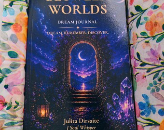 May include: A rectangular dream journal titled Between Worlds by Julita Dirsaite J Soul Whisper, featuring a dark blue cover with a stone archway, crescent moon, and crystals, nestled inside a cardboard box lined with floral tissue paper.