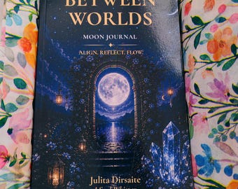 May include: A dark blue journal titled Between Worlds Moon Journal by Julita Dirsaite J Soul-Whisper, featuring a full moon, stone archway, and crystals, nestled in a cardboard box lined with white tissue paper printed with colorful flowers.