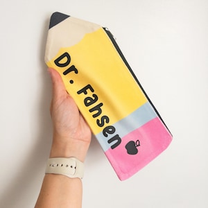 May include: A pencil-shaped pencil case in yellow, black, and pink, with the text "Dr. Fahsen" printed on it. The pencil case has a black zipper and a small apple graphic.