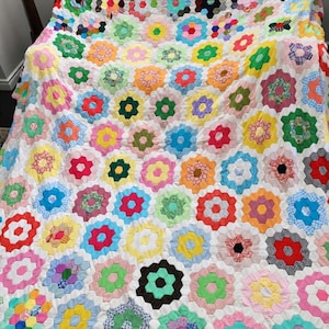 Könnte beinhalten: Ein handgefertigter Quilt mit einem sich wiederholenden Muster aus mehrfarbigen Stoffsechsecken, die in blumenartigen Clustern angeordnet sind. Der Quilt ist mit weißen Stoffrändern zwischen jedem Sechseck konstruiert und zeigt eine Vielzahl von Drucken und Unifarben.