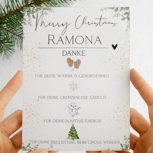 Könnte beinhalten: Eine Weihnachtskarte mit den Worten "Merry Christmas Ramona" in eleganter Schrift. Die Karte enthält das Wort "DANKE" und mehrere Sätze auf Deutsch. Dekorative Elemente sind ein Herz, Fäustlinge, ein Schneeflocke und ein Schneemann.