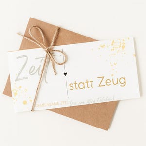 以下が含まれることがあります： 「Zeit statt Zeug」の文字が銀色と金色で書かれた白いカードが、茶色の封筒に紐で結ばれています。カードには金色のペイントスプラッシュと小さな黒いハートがあります。