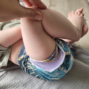 Könnte beinhalten: Ein Baby trägt eine blaue und violette gemusterte Windelhülle mit weißem Besatz. Die Windelhülle hat ein Blumenmuster mit goldenen Akzenten.