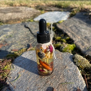 May include: A clear glass bottle with a black dropper top, filled with a golden liquid and botanicals. The bottle is adorned with a small heart charm, a pink gemstone, and tied with twine. The bottle is sitting on a stone surface.