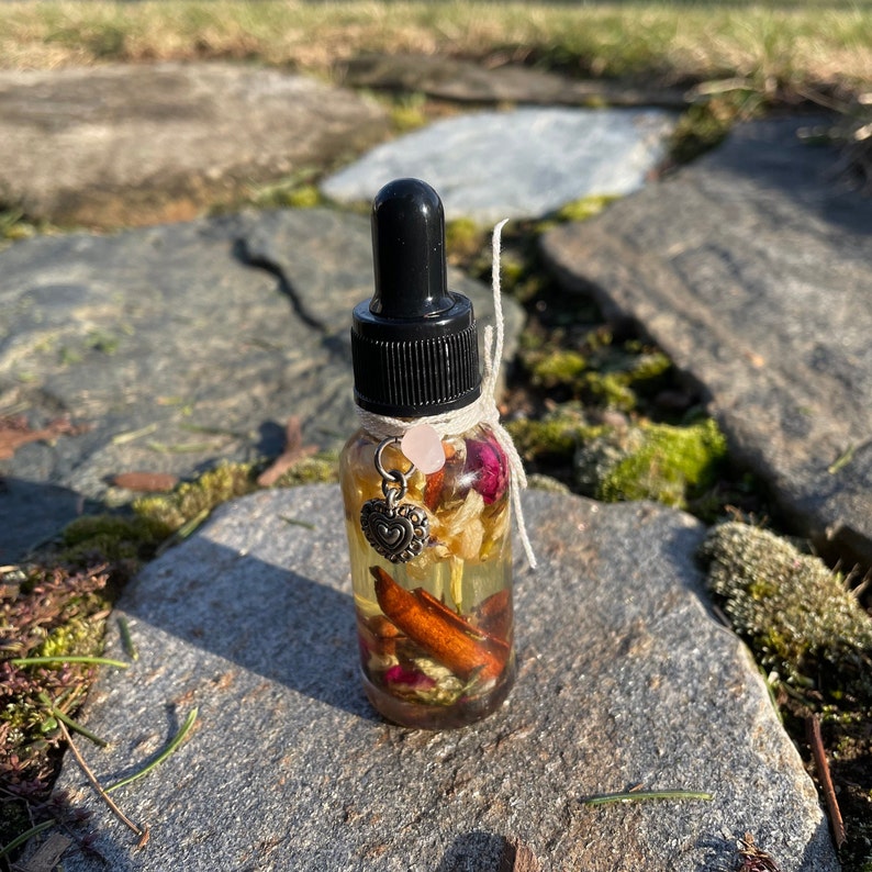 May include: A clear glass bottle with a black dropper top, filled with a golden liquid and botanicals. The bottle is adorned with a small heart charm, a pink gemstone, and tied with twine. The bottle is sitting on a stone surface.