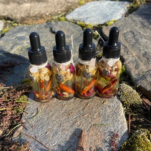 May include: Four small glass dropper bottles filled with a golden liquid, herbs, and spices. Each bottle has a black dropper top, a twine tie, and a heart charm with a pink crystal. The bottles are arranged on a stone surface.