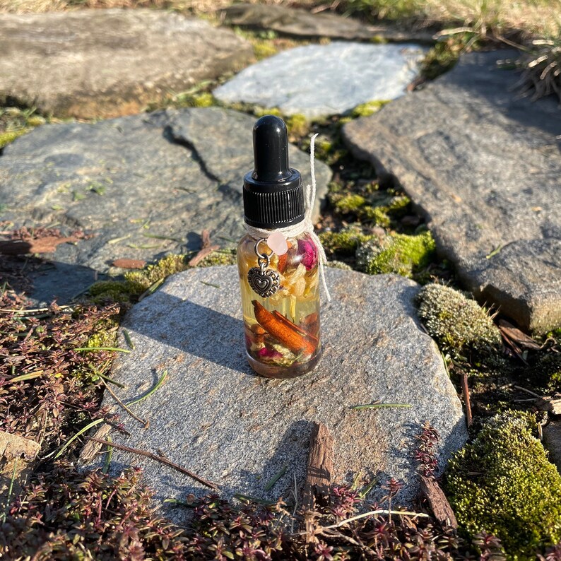 May include: A clear glass essential oil bottle with a black dropper top. The bottle contains dried flowers, cinnamon sticks, and a heart charm. A pink gemstone and a white string are tied around the neck of the bottle.