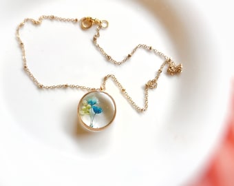 May include: A gold chain necklace with a round pendant featuring pressed blue and white flowers encased in clear resin.