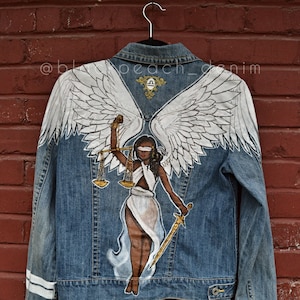 May include: A blue denim jacket with white painted wings and a figure of a blindfolded woman holding scales and a sword. The woman is painted in brown, white, and gold. The text "@blackpeach_denim" is visible on the jacket.