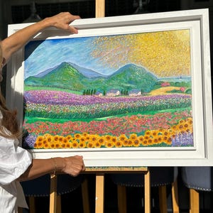 May include: A framed landscape painting depicting a vibrant field of sunflowers, lavender, and other colorful flowers. Green mountains and small houses are in the background under a bright yellow and blue sky. The painting is in a white frame.