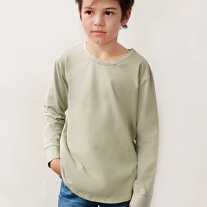 May include: A person wearing a long-sleeved, pale green top and blue jeans. The top has a crew neck and cuffed sleeves. The person is standing with one hand in their pocket. The background is plain.