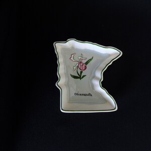 May include: A white ceramic dish shaped like the state of Minnesota. The dish is decorated with a green outline and a pink and green wildflower. The word "Minnesota" is written in black at the bottom of the dish.