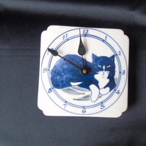 May include: Square wall clock with a white face and a blue cat illustration. The clock face has black hands and blue numerals. The cat is depicted in blue with white accents. The clock is a decorative item.