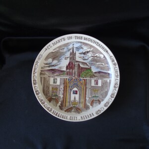Può includere: Un piatto in ceramica bianca con un'illustrazione marrone e bianca di St. Mary's in the Mountains, Virginia City, Nevada. L'illustrazione rappresenta una chiesa con un campanile e un paesaggio circostante.