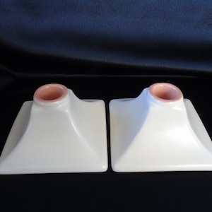 May include: A pair of white ceramic candle holders with a square base and a pink center. The holders are designed to hold a standard-sized candle.