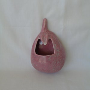 May include: A pink ceramic wall pocket shaped like an eggplant. The wall pocket has a speckled finish with light green accents and a small opening at the top. The interior is a darker shade of pink.