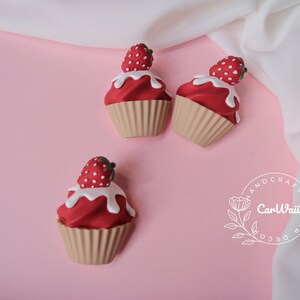 May include: Three miniature red and white cupcake decorations with strawberry toppers. The cupcakes are on a pink background.