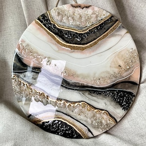 Könnte beinhalten: Runde Wandkunst aus Harz mit einem abstrakten Design in Weiß, Schwarz und Gold. Das Design verfügt über wirbelnde Linien und einen geschichteten Effekt, der an Achat oder Marmor erinnert.
