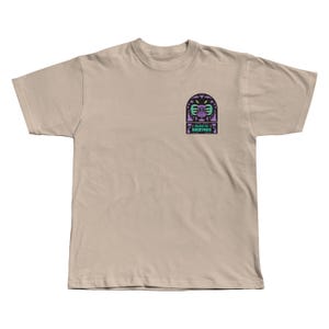 Puede incluir: Camiseta color beige con un dise&ntilde;o gr&aacute;fico en el pecho izquierdo. El dise&ntilde;o incluye una canasta de disc golf estilizada, palmeras y el texto "I BEAT THE BOGEYMAN". El dise&ntilde;o utiliza colores morados y turquesas. Una camiseta casual.