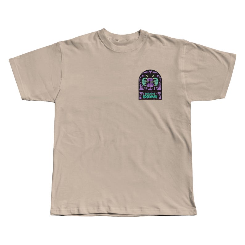 Puede incluir: Camiseta color beige con un dise&ntilde;o gr&aacute;fico en el pecho izquierdo. El dise&ntilde;o incluye una canasta de disc golf estilizada, palmeras y el texto "I BEAT THE BOGEYMAN". El dise&ntilde;o utiliza colores morados y turquesas. Una camiseta casual.