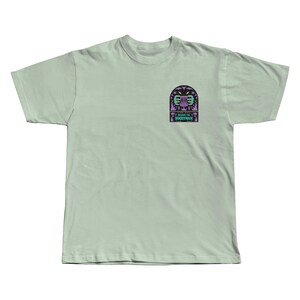 Puede incluir: Una camiseta verde claro con un peque&ntilde;o gr&aacute;fico en el pecho izquierdo. El gr&aacute;fico presenta una canasta de disc golf estilizada con un dise&ntilde;o p&uacute;rpura y verde, y el texto "I BEAT THE BOGEYMAN".