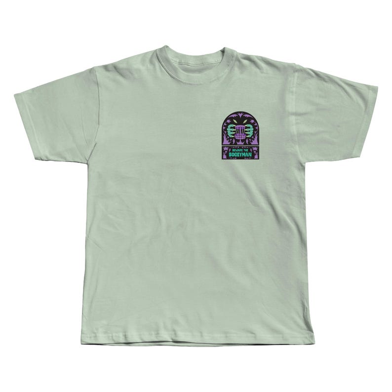 Puede incluir: Una camiseta verde claro con un peque&ntilde;o gr&aacute;fico en el pecho izquierdo. El gr&aacute;fico presenta una canasta de disc golf estilizada con un dise&ntilde;o p&uacute;rpura y verde, y el texto "I BEAT THE BOGEYMAN".