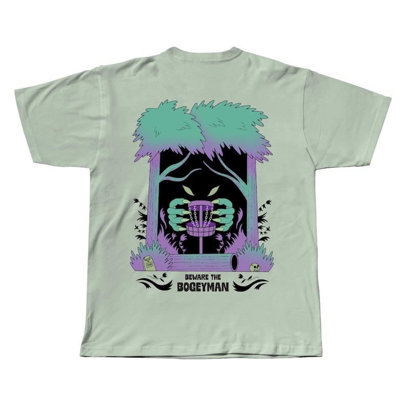 Puede incluir: Camiseta verde claro con un gr&aacute;fico de una canasta de disc golf con manos y ojos de monstruo, enmarcada por &aacute;rboles. El texto "BEWARE THE BOGEYMAN" est&aacute; debajo. El dise&ntilde;o utiliza morado, verde y negro.