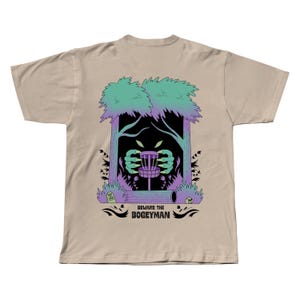 Puede incluir: Camiseta color beige con un dise&ntilde;o gr&aacute;fico. El dise&ntilde;o presenta una canasta de disc golf con manos de monstruo que la alcanzan, &aacute;rboles, arbustos y el texto "Beware the Bogeyman" en negro.