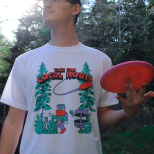May include: A white t-shirt with a graphic of a cartoon character holding a disc golf basket. The graphic says "Take the Local Route". The person is holding a pink disc golf disc.